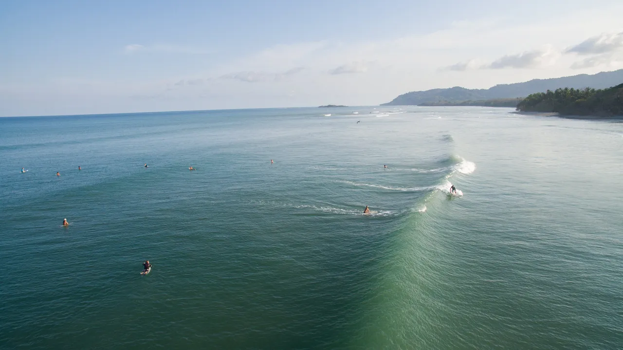 Surfers waiting in a wide green lineup, seen from above.