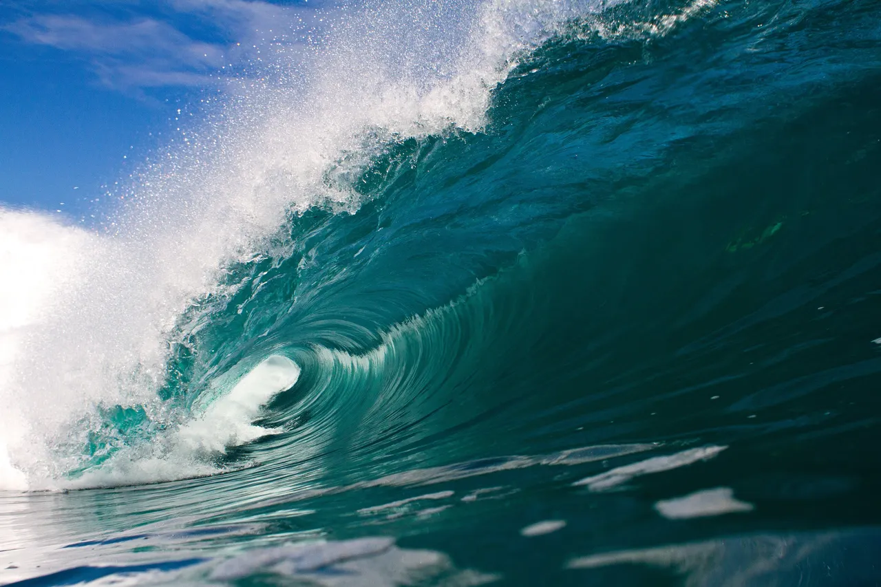 A turquoise wave curling into a barrel under a blue sky.