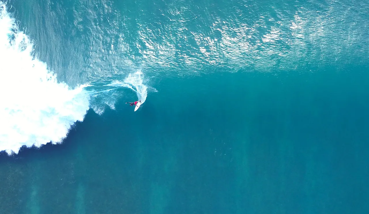 A surfer carving across a blue wave, seen from directly above.