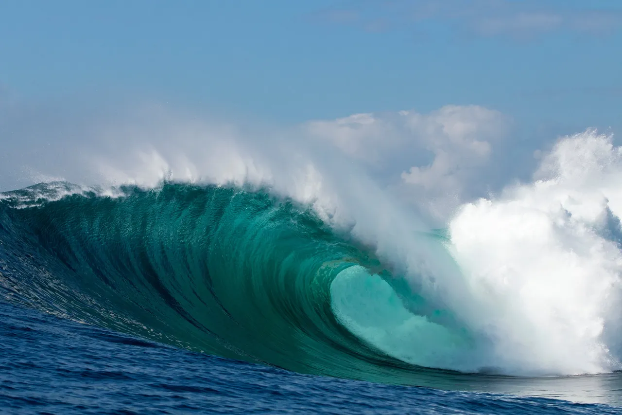 A large green wave pitching over, white spray blowing back into the sky.