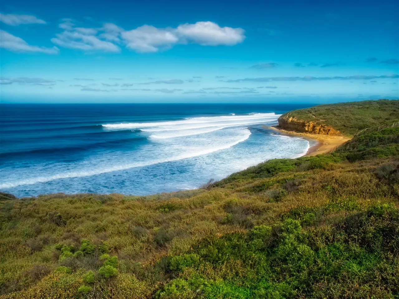 Long lines of swell rolling into a curved bay below green coastal hills.